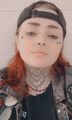 Me with a black hat, bridge and septum ring, black shirt, denim jattle jacket, shoulder length red hair, and snapchat neck tat filter